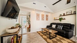 A modern living room with a black leather sofa, a wooden coffee table, a flat-screen TV mounted on the wall, and a patterned rug. - NEW! Boho Bungalow (Carolina Beach)