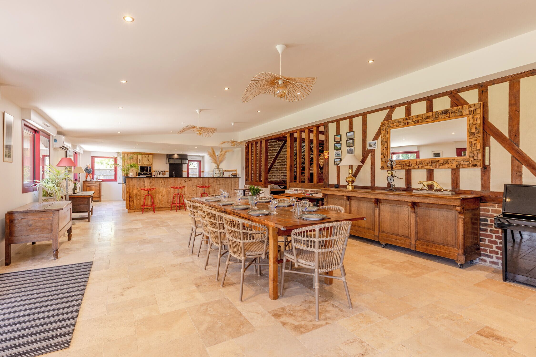 A spacious dining area with a long wooden table and wicker chairs, a kitchen with a brick wall and wooden beams, and a staircase leading to an upper level.
