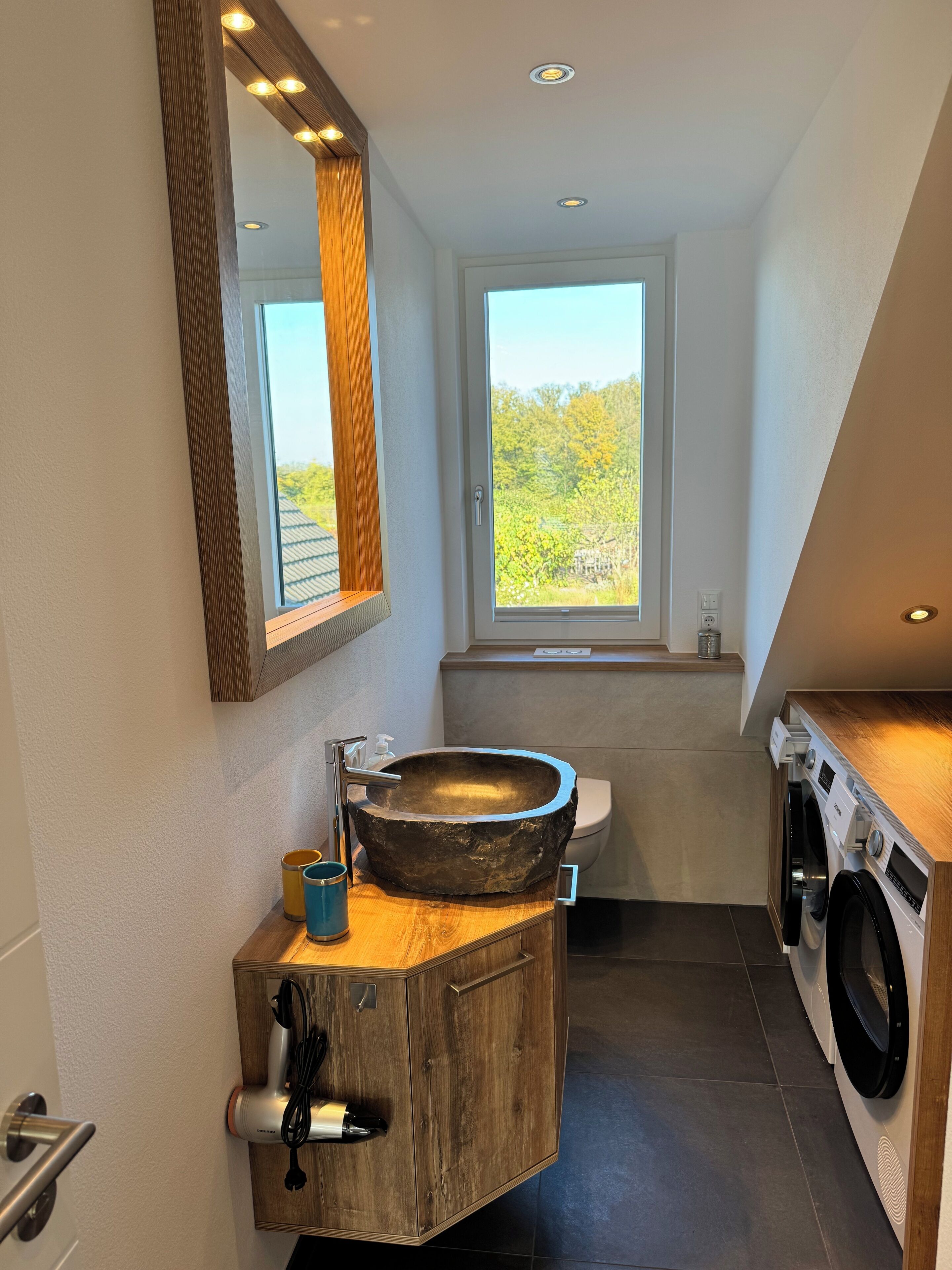 A modern bathroom with a large wooden vanity, a vessel sink, a built-in washing machine, and a window with a view of greenery.