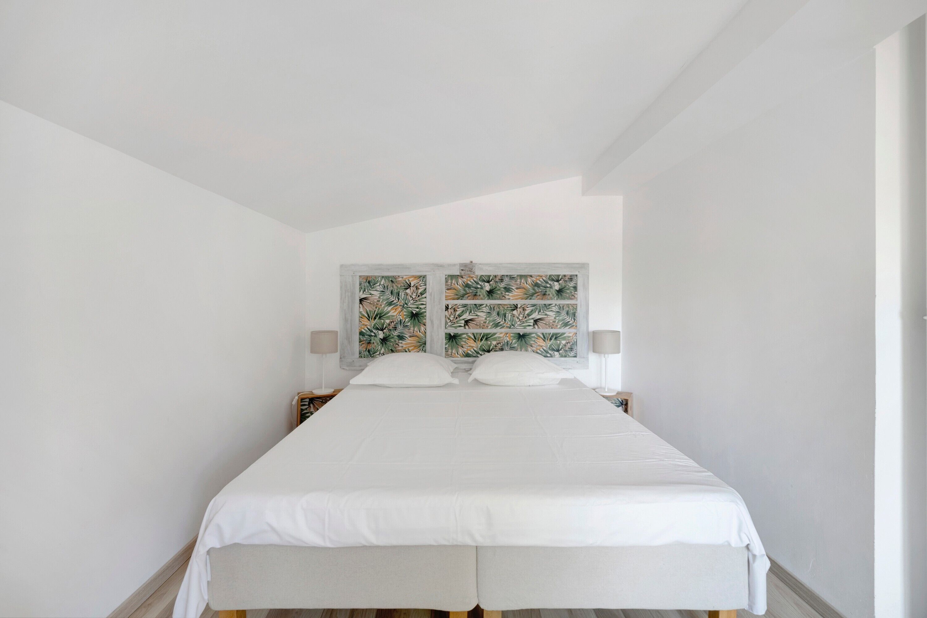 Quarto minimalista com uma cama de casal, roupa de cama branca e cabeceira decorativa com padrão de folha tropical.