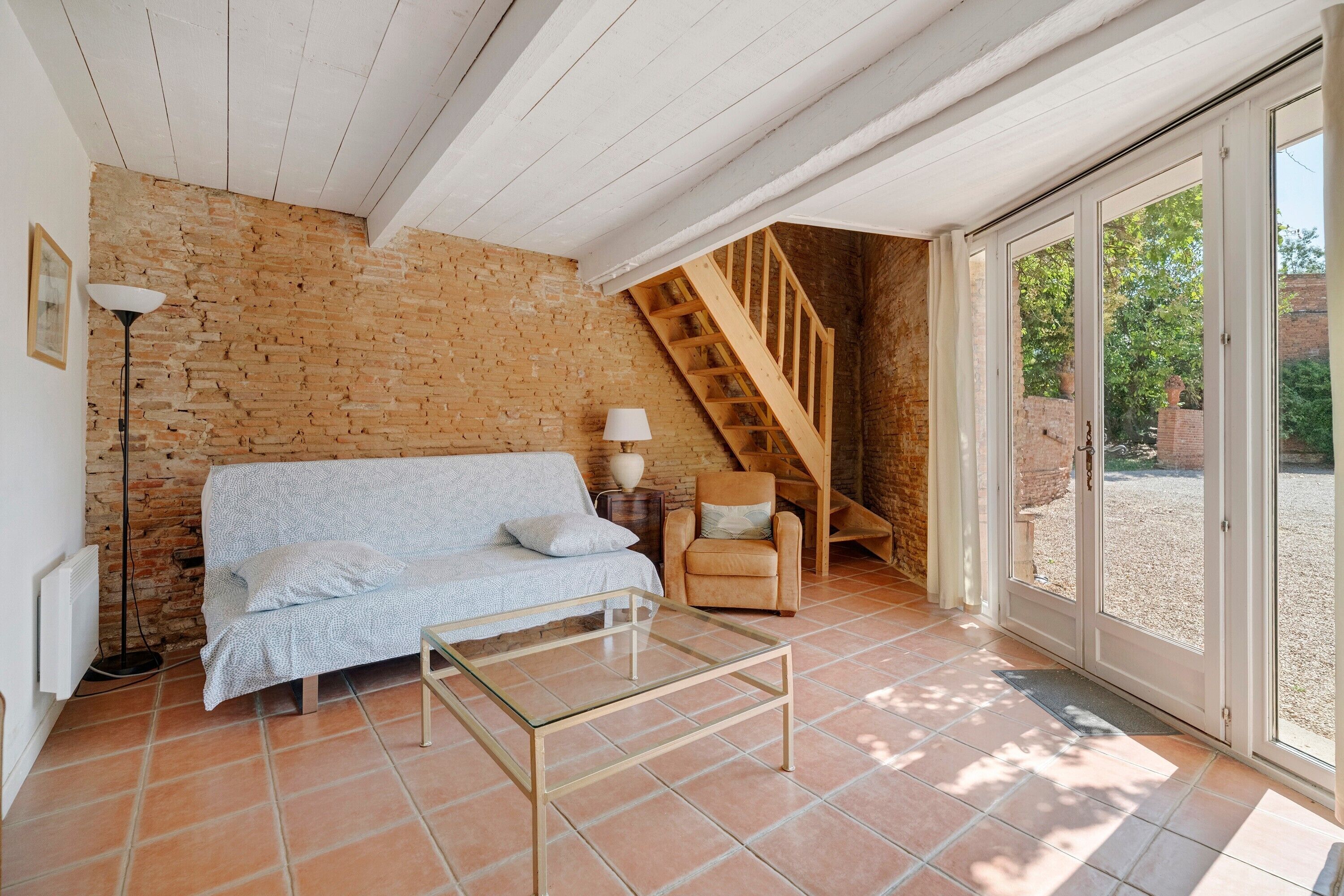 A room with a brick wall, a wooden staircase, a sofa, a chair, and a glass table.