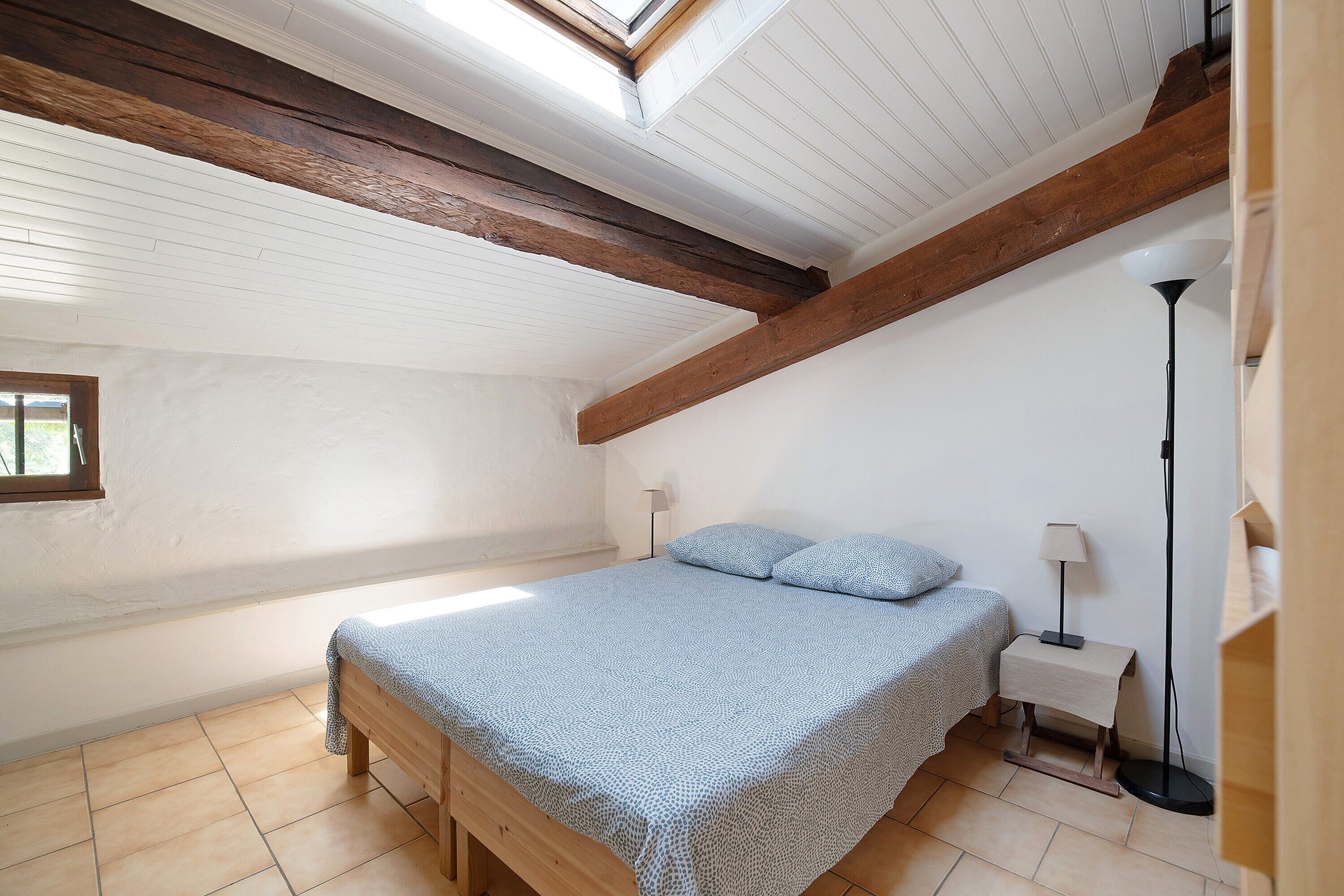 A bedroom with a sloped ceiling, wooden beams, a bed with a patterned bedspread, a bedside table with a lamp, and a floor lamp.