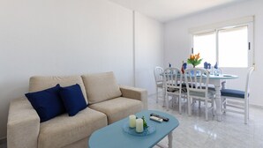 A living room with a beige sofa, blue cushions, a round table with chairs, and a window with blinds. - Bohemian Nights by Fidalsa (Torrevieja)