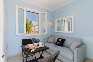 Une pièce avec un mur bleu clair, un canapé gris, une table noire avec des livres et une tasse blanche, et une fenêtre dont le volet est ouvert.