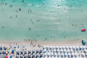 An aerial view of a beach with people swimming and sunbathing, and rows of umbrellas and lounge chairs along the shore. - Mommy House by BarbarHouse (Torre Lapillo)