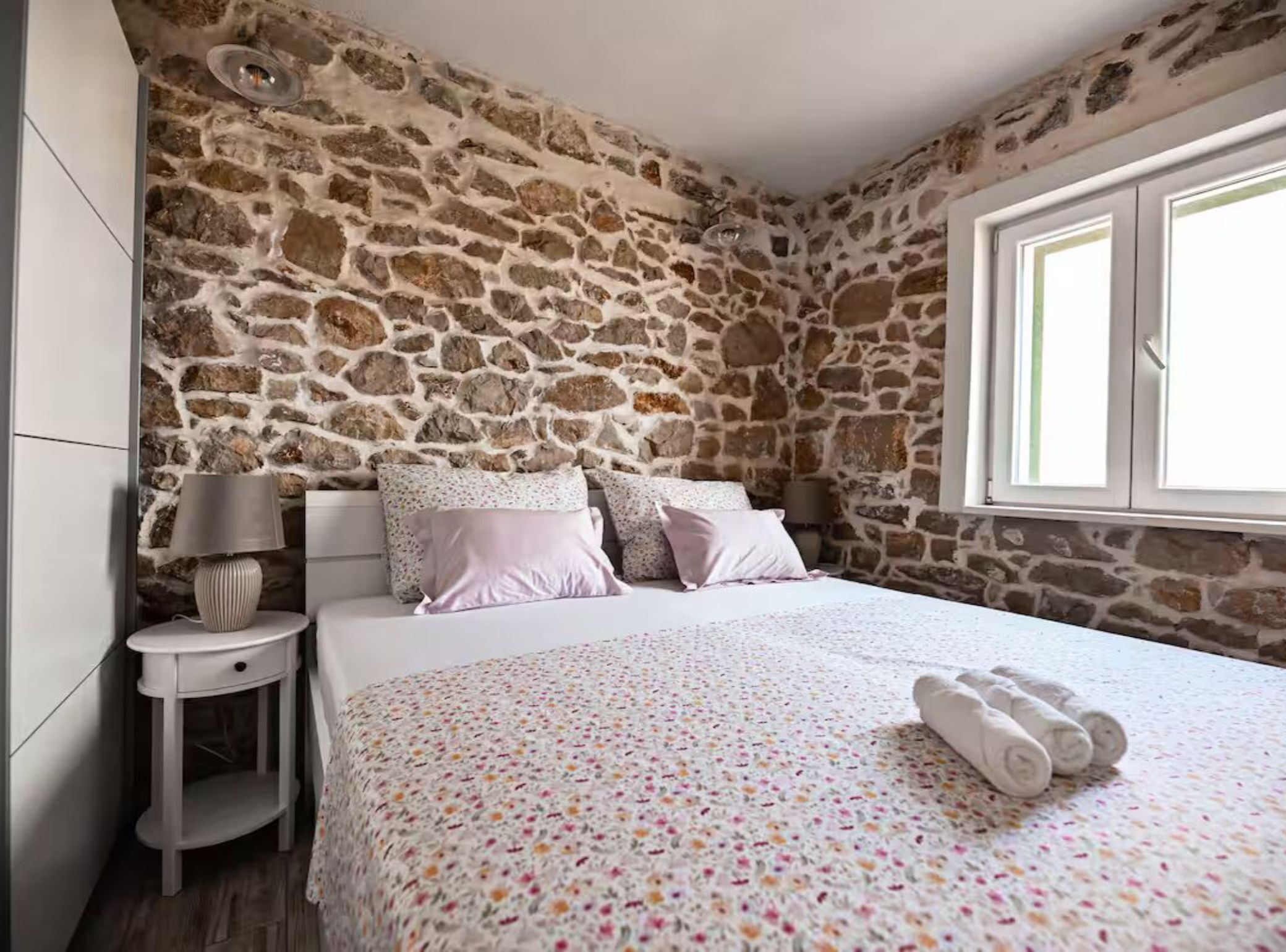 Un dormitorio con pared de piedra, una cama con ropa de cama floral, una mesita de noche con lámpara y una ventana.
