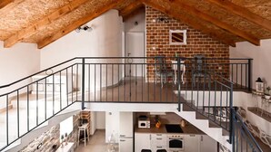 Ein Loft-Zimmer mit freiliegenden Holzbalken, einer Ziegelwand, einem Balkon mit Metallgeländer und einer Küchenzeile mit weißen Schränken und Geräten.