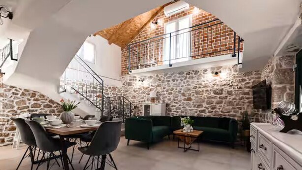 Ein Loftsitz mit Steinwand, einem Essbereich mit Stühlen, einem Sofa und einer Treppe zu einer Erker Ebene.