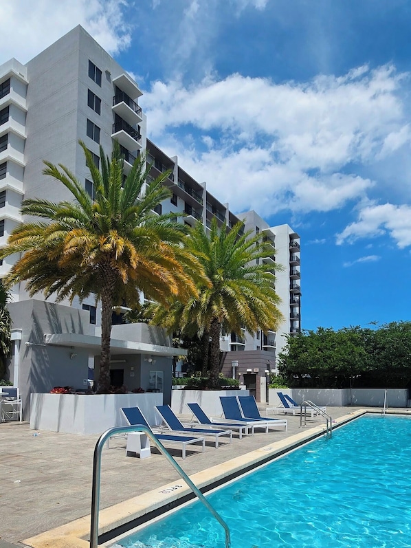 A swimming pool with lounge chairs, a palm tree, and a modern building with balconies.