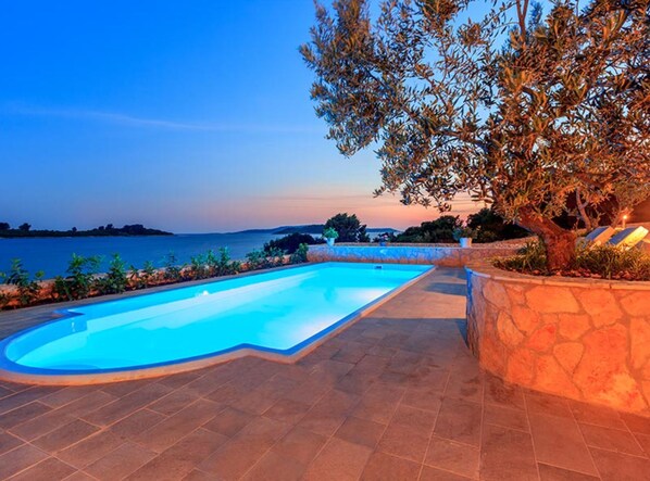 A swimming pool with a stone border, a view of the sea, and a tree with illuminated leaves. - Villa Renata (Okrug)