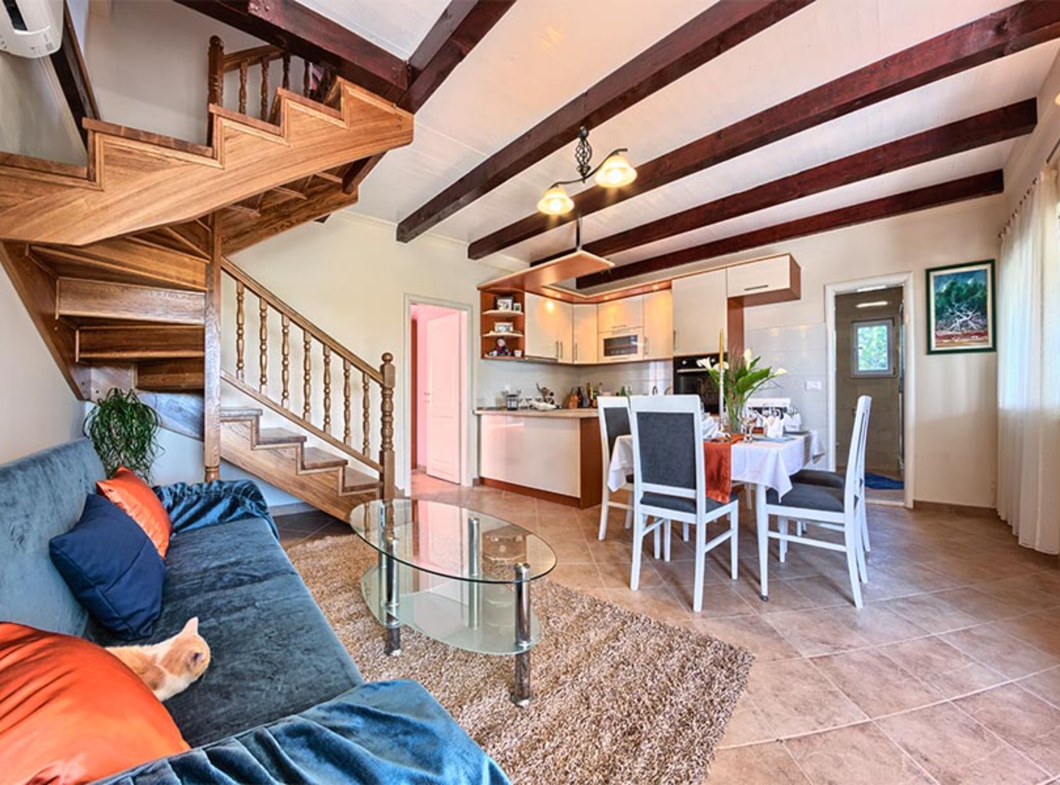 A living room with a glass coffee table, a sofa with cushions, a staircase, a dining area with chairs, and a kitchen in the background.