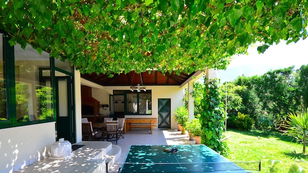 Uma área de piscina ao ar livre com mesa e cadeiras, rodeada por vegetação e um espaço de estar coberto.