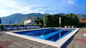 Piscina com espreguiçadeiras e guarda-sóis, um edifício com telhado vermelho e montanhas ao fundo.