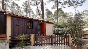 A rustic wooden cabin with a tiled roof, surrounded by a gravel path and a wooden fence. - Holiday Home "Petite Semi-Detached Villa in the Pine Forest" with Wi-Fi (Lacanau)