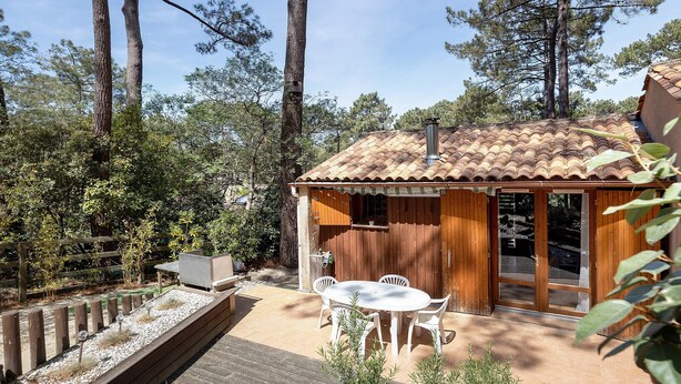 A wooden cabin with a tiled roof, a patio with a table and chairs, and a wooden deck. - Holiday Home "Petite Semi-Detached Villa in the Pine Forest" with Wi-Fi (Lacanau)