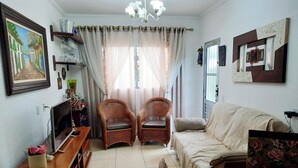 A living room with a sofa, two wicker chairs, a TV on a wooden stand, a chandelier, and a window with sheer curtains.
