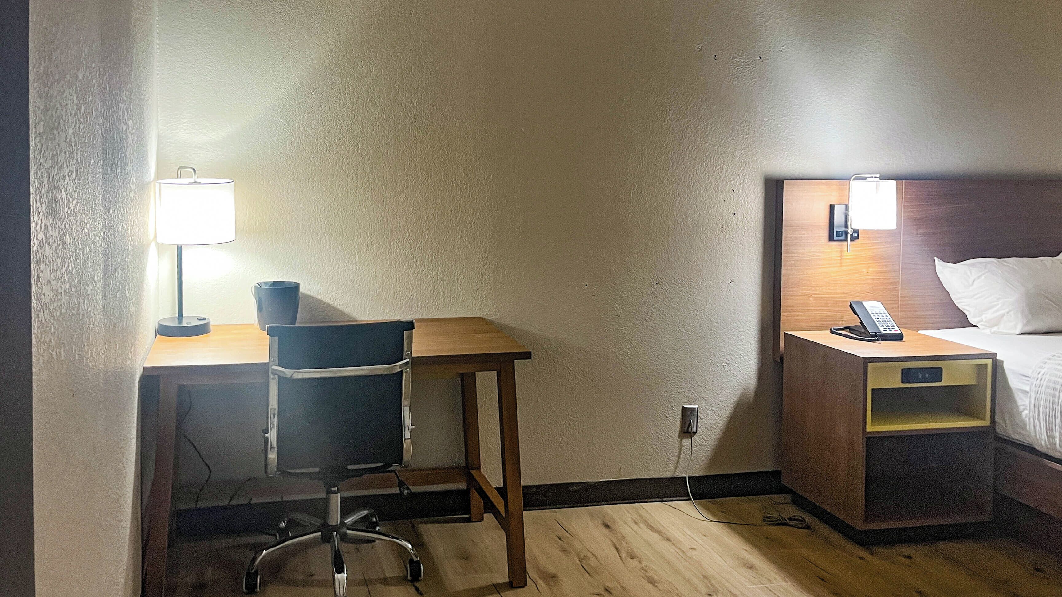 Quarto de hotel com escrivaninha, cadeira, luminária, mesa de cabeceira com telefone e cama.