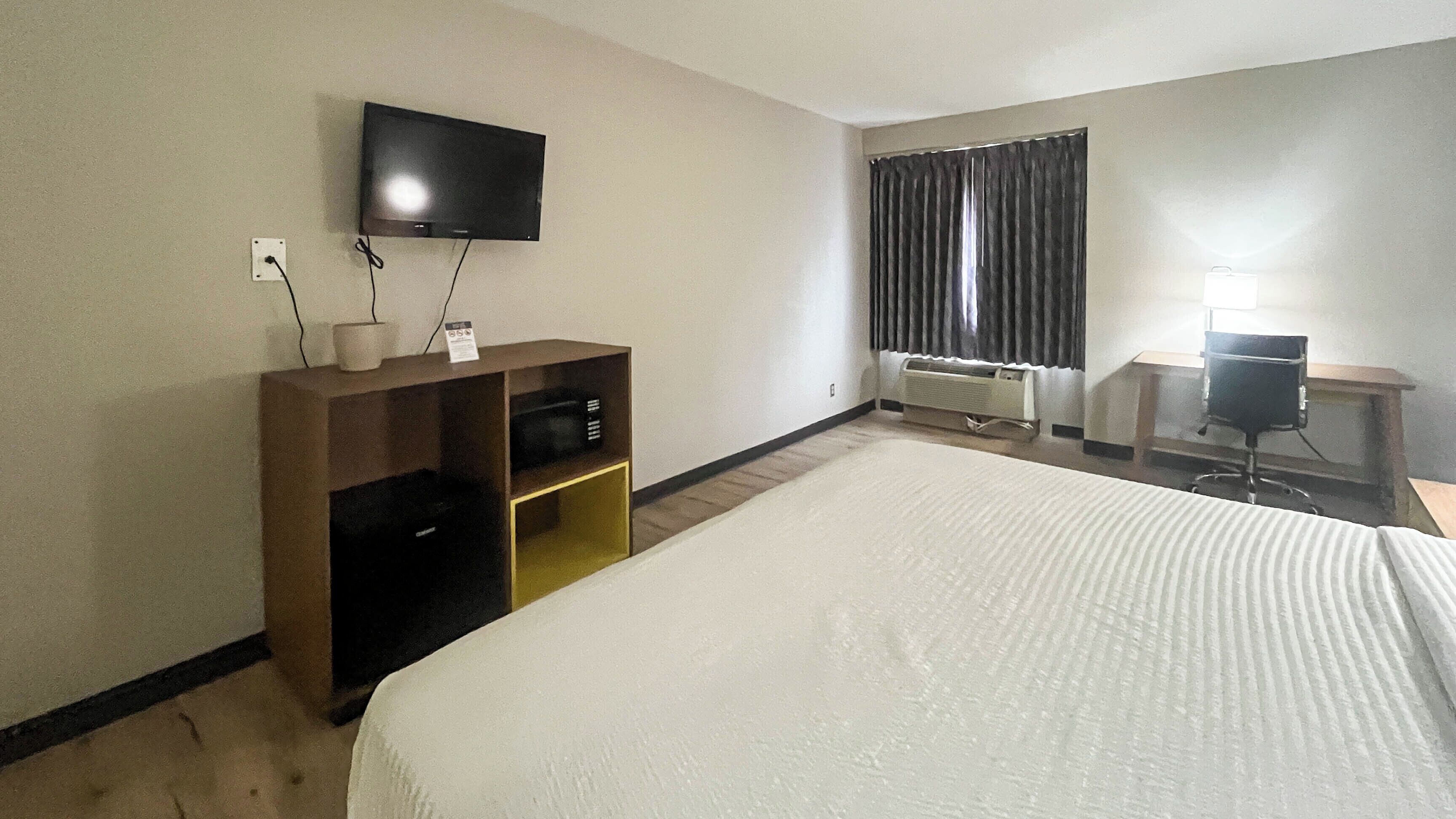 A hotel room with a bed, a TV mounted on the wall, a desk with a chair, and a window with curtains.