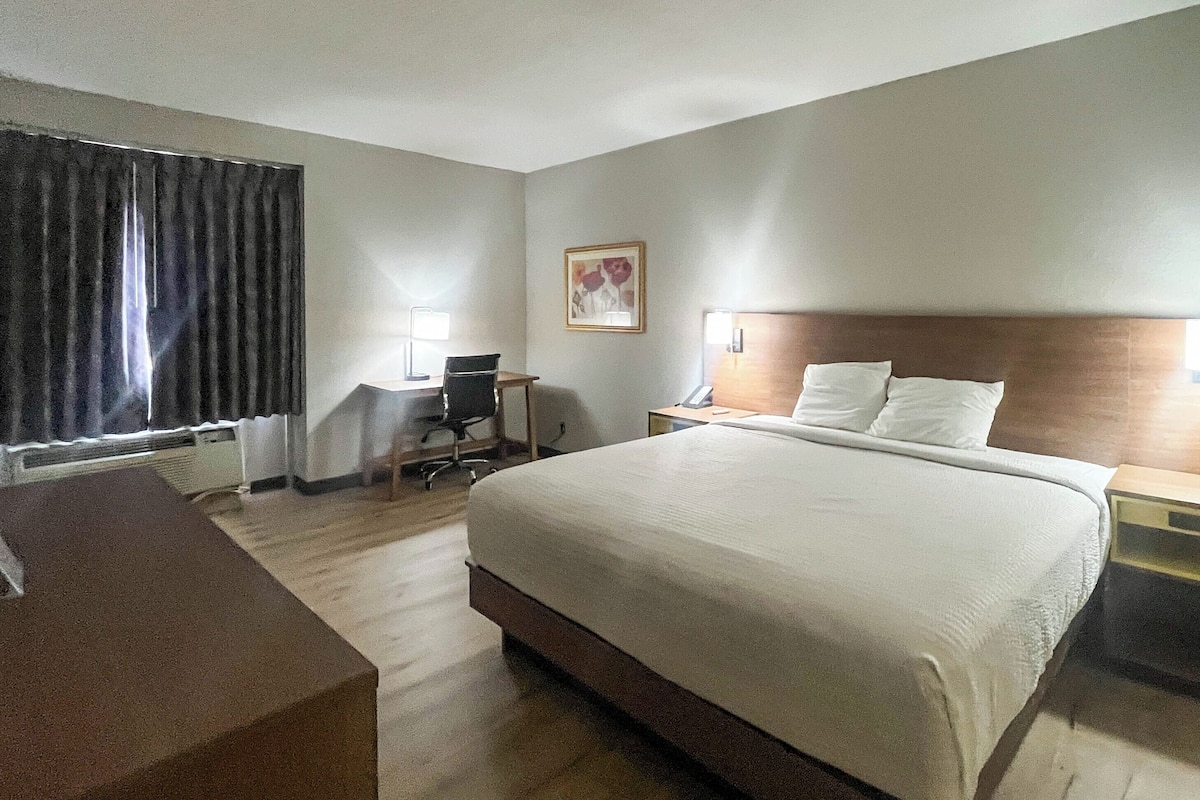 A hotel room with a large bed, a desk, a chair, a nightstand, and a window with curtains.