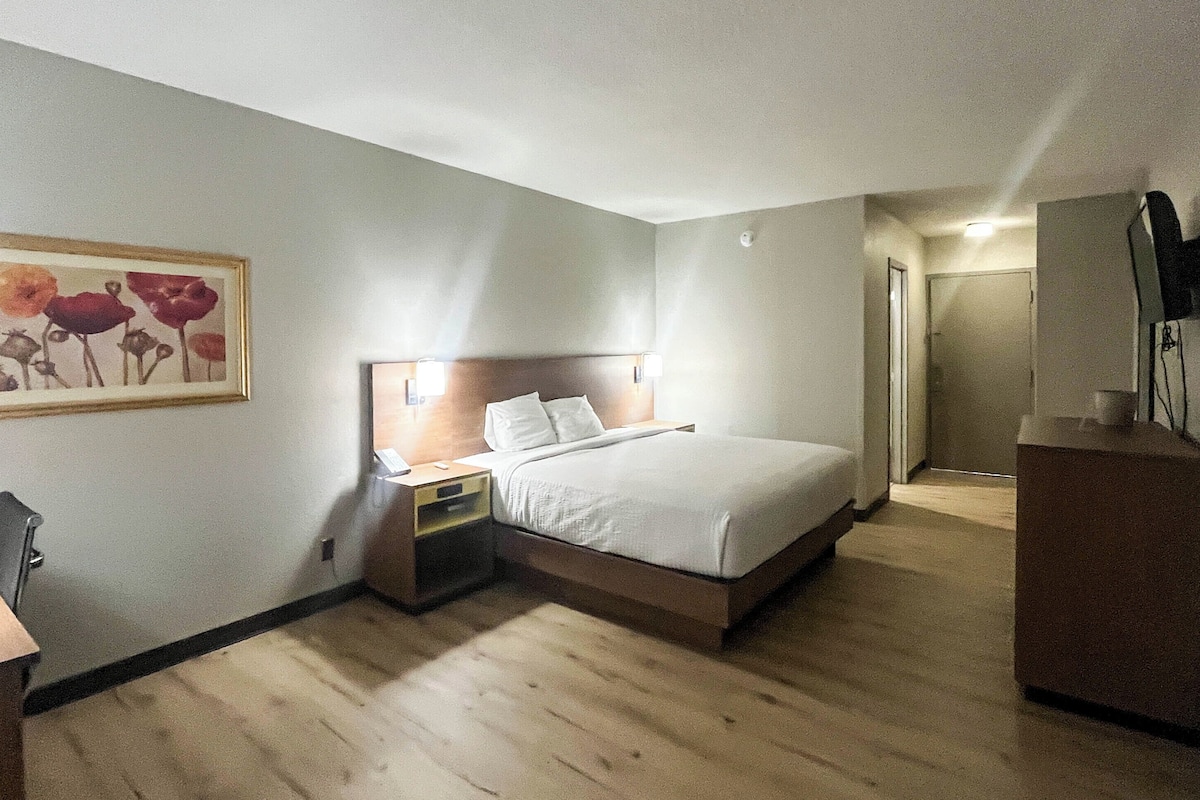 A hotel room with a bed, bedside table, chair, desk, and a painting on the wall.