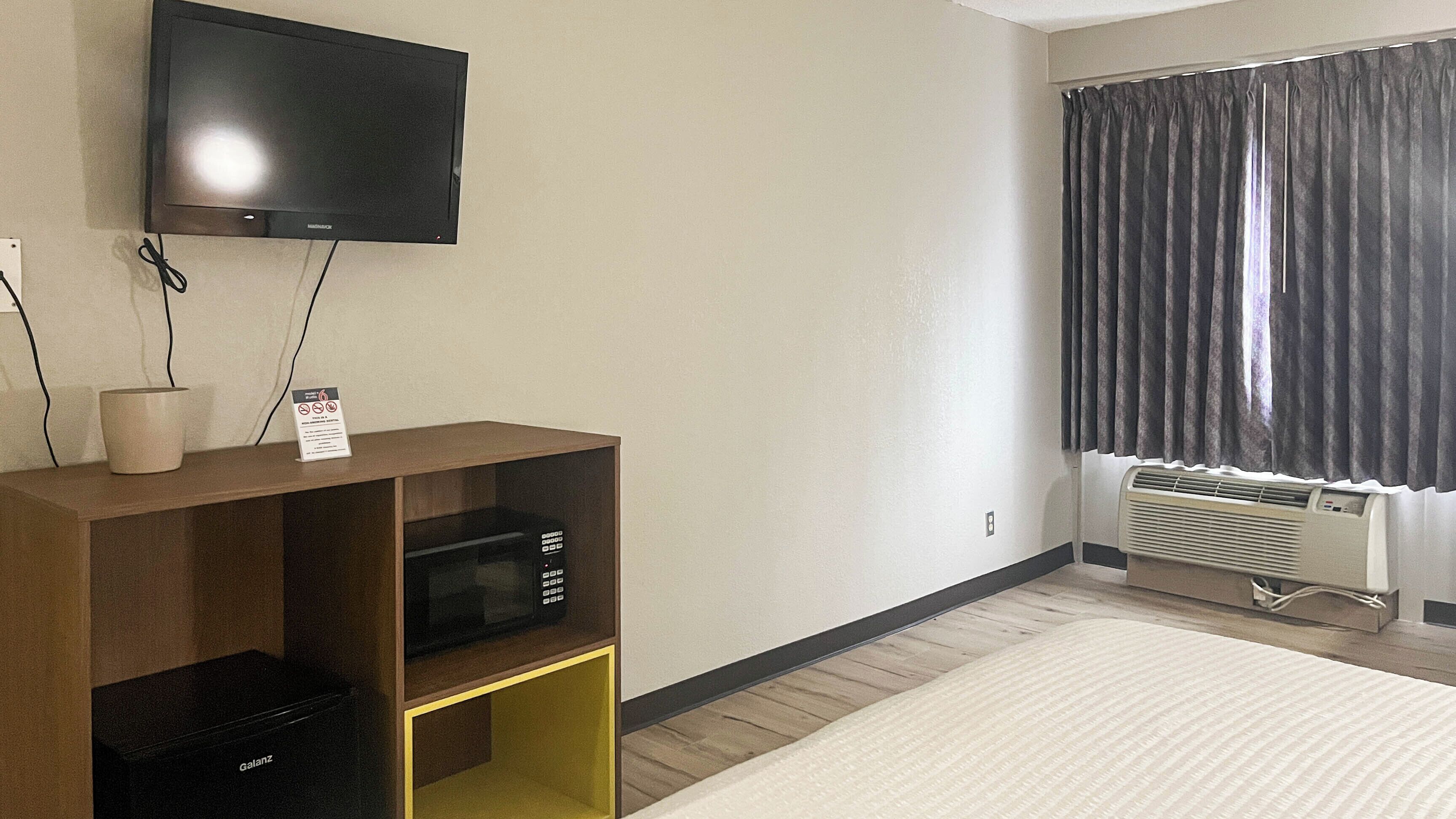 A hotel room with a flat-screen TV mounted on the wall, a wooden entertainment unit with a microwave, a small table with a cup, a window with dark curtains, and a radiator.