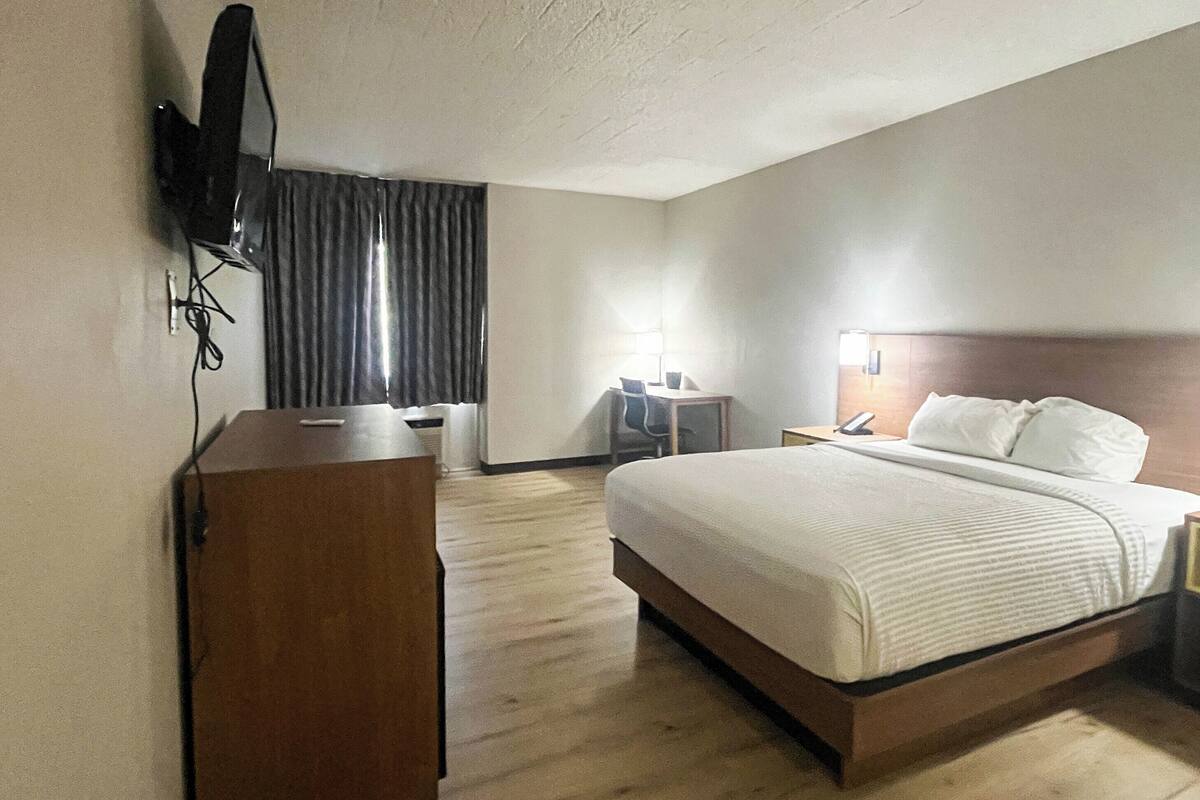 A hotel room with a bed, bedside tables, a desk, a chair, a TV mounted on the wall, and a window with curtains.