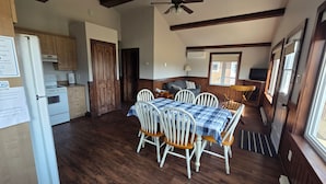 A room with a wooden floor, a dining table with chairs, a sofa, a ceiling fan, and a kitchen area with a refrigerator and stove. - Motel & Chalets l'Acadien sur mer (Petit-Rocher)
