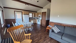 A living room with a sofa and wooden chairs, a dining area with a table and chairs, and a kitchen with appliances. - Motel & Chalets l'Acadien sur mer (Petit-Rocher)