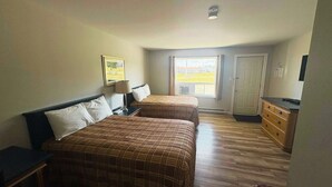 A hotel room with two beds, a dresser, a window with a view, and a ceiling light. - Motel & Chalets l'Acadien sur mer (Petit-Rocher)