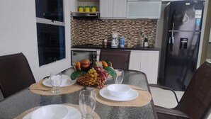 A dining table set with plates, glasses, and a fruit basket, with a kitchen in the background.