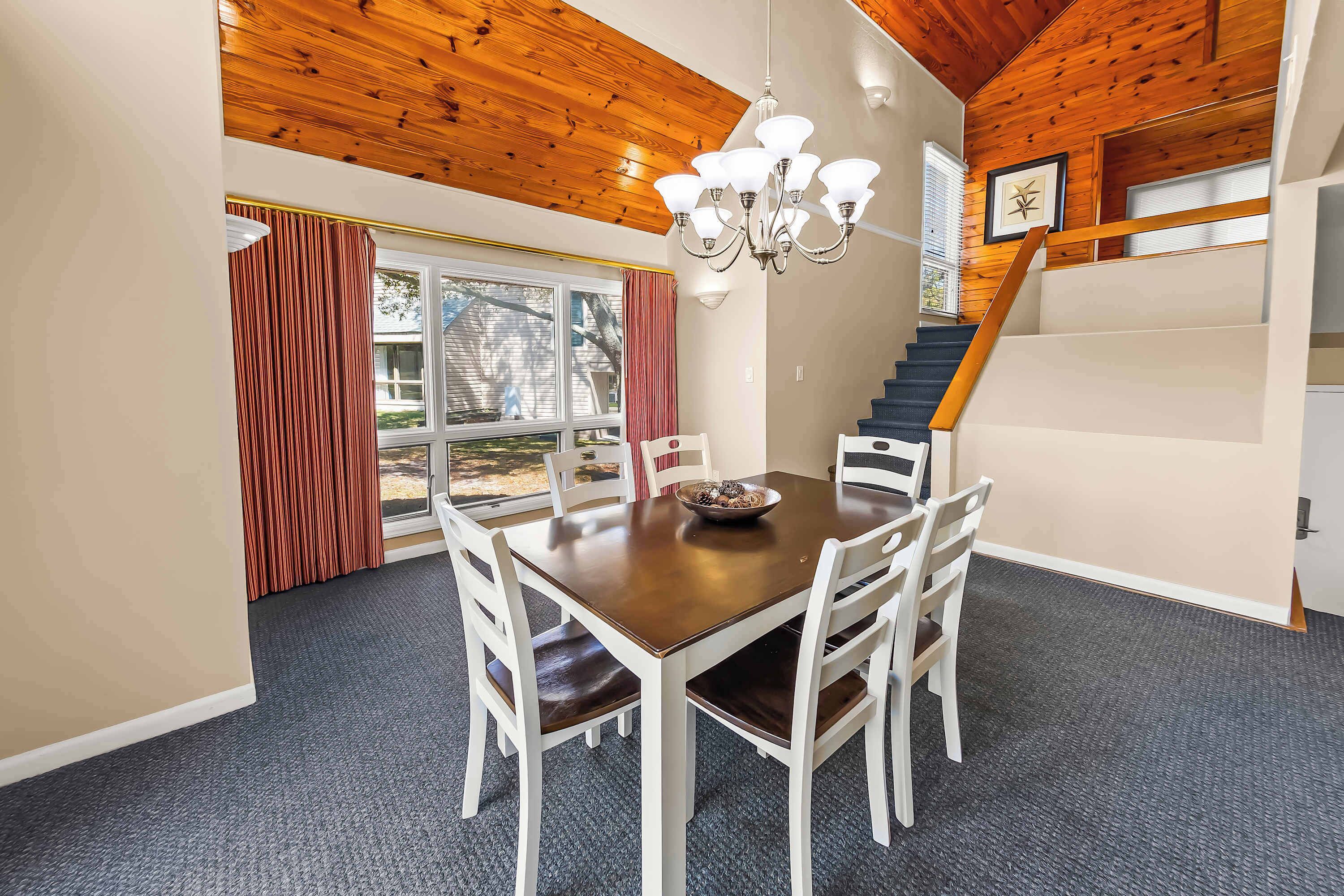 Un espace repas comprenant une table en bois, des chaises blanches, un lustre et un escalier.
