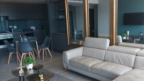 Moderni olohuone, jossa on sohva, ruokailutila tuoleineen, keittiö liesineen sekä seinälle asennettu televisio.