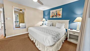 A hotel room with a bed, a ceiling fan, a nightstand with a lamp, a painting on the wall, and a mirror.
