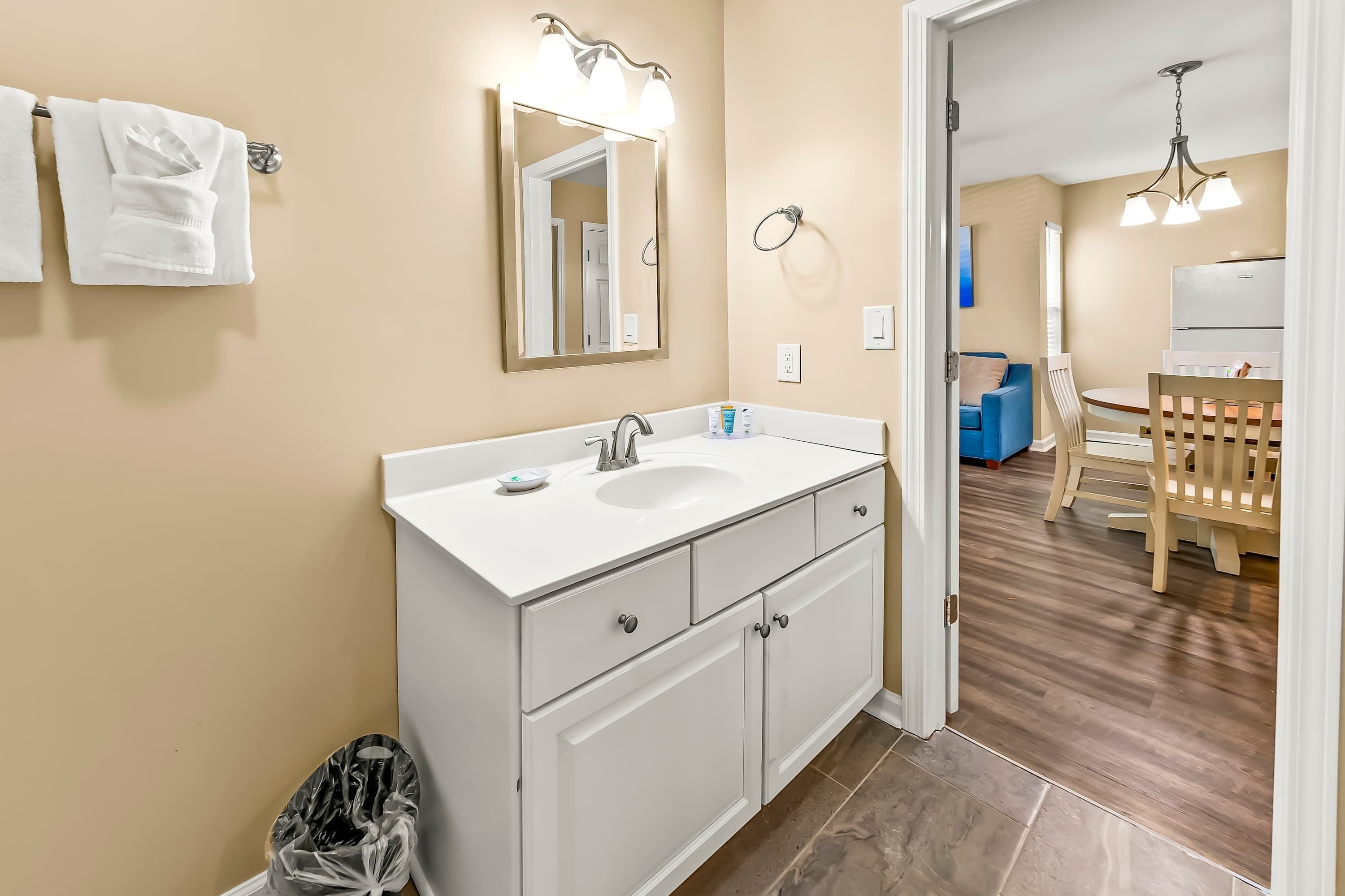 A bathroom with a white sink, mirror, and cabinets. There is a trash bin and a towel rack with towels.