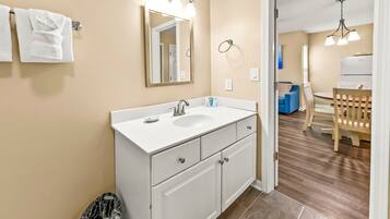 A bathroom with a white sink, mirror, and cabinets. There is a trash bin and a towel rack with towels.