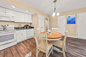 A kitchen with white cabinets, a microwave, a stove, and a dining area with a wooden table and chairs. - Family Friendly Condo with Golf, Pools & River Access! (New Bern)