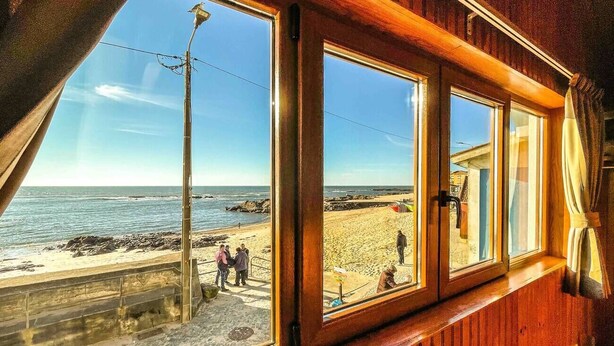 Una habitación con amplios ventanales que dan a una playa, paredes de madera y vistas al mar y a personas en la playa.