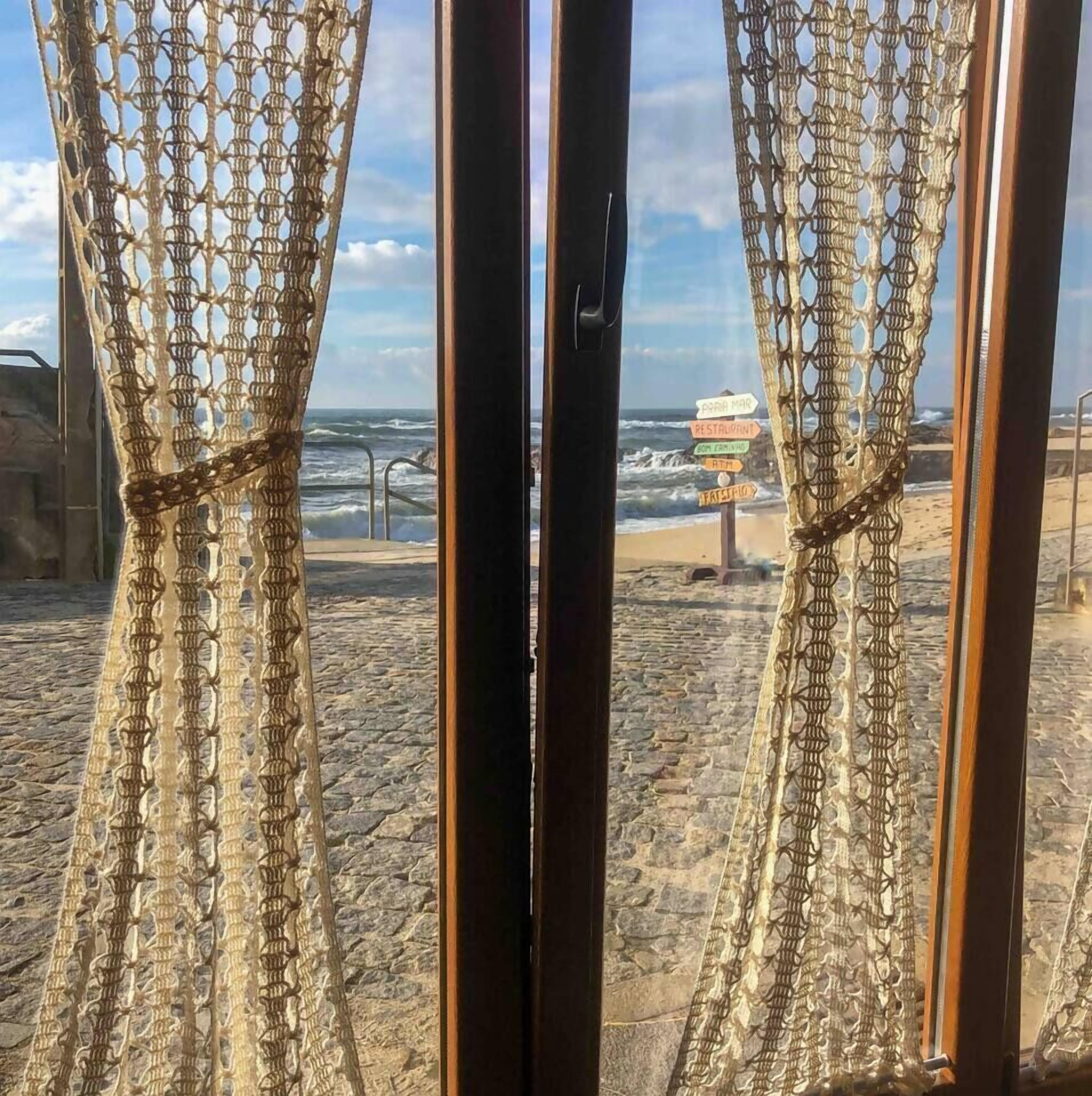 A beach view through a window with a decorative curtain, a signpost with directions, and a cobblestone path.