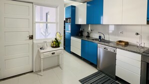 A modern kitchen with white and blue cabinets, stainless steel appliances, and a small white table with a vase of flowers. - Ninho do Carriço - Beach 200m away - Patio & KidsZone (Vila Chã)