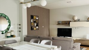 A modern living room with a dining area, a sofa, a TV, and a decorative wall panel. - Ninho do Carriço - Beach 200m away - Patio & KidsZone (Vila Chã)