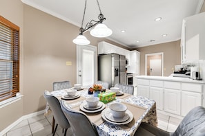 A dining area with a table set for four, a kitchen in the background, and a window with blinds. - Warm & Spacious 4BR/3BA/4TV Home – Newly Renovated (Forney)