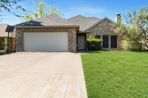 A two-story house with a brick exterior, a white garage door, and a well-maintained lawn. - Warm & Spacious 4BR/3BA/4TV Home – Newly Renovated (Forney)
