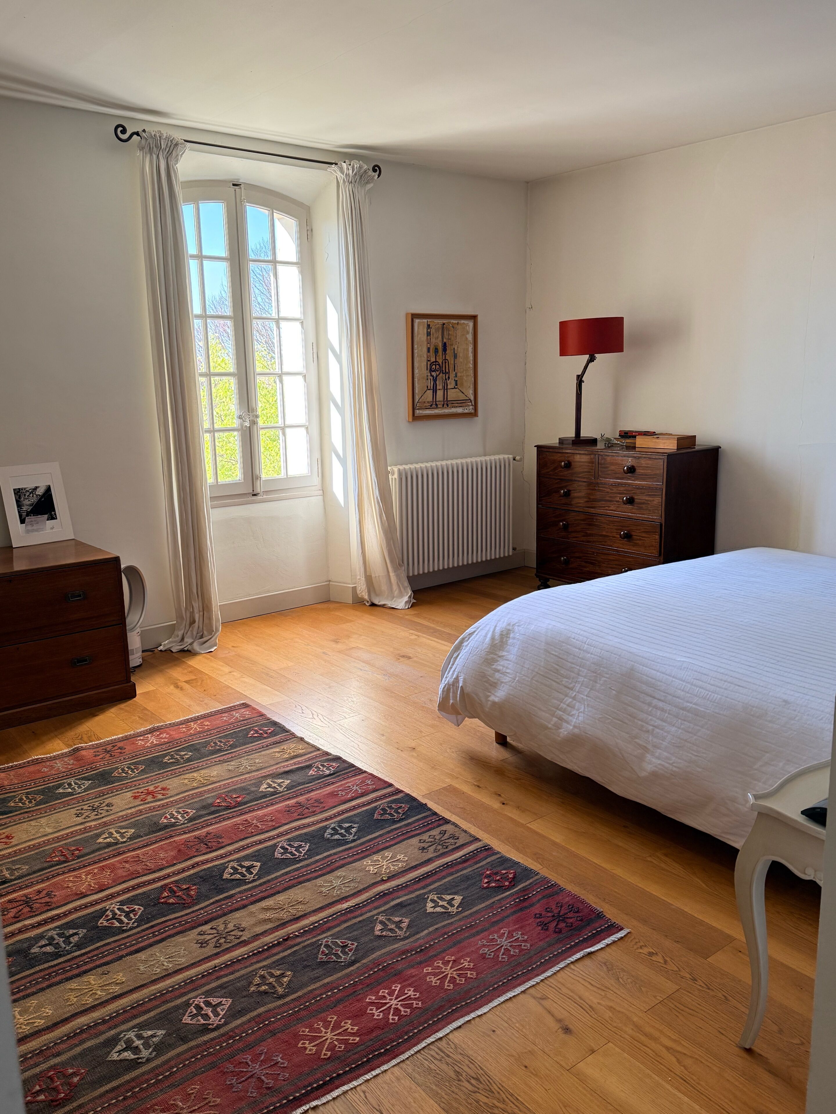 Une chambre à coucher avec un lit, une commode, un radiateur, une fenêtre avec des rideaux, un tapis et un tableau accroché au mur.