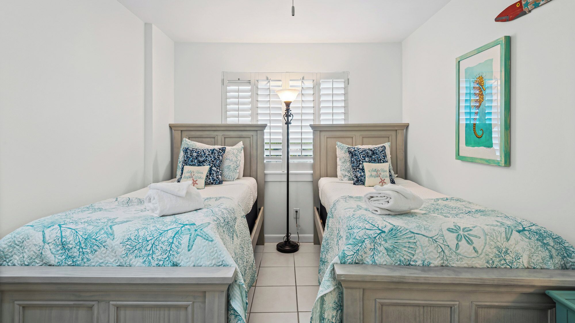 A bedroom with two beds, each with a floral bedspread, a floor lamp, and a framed seahorse artwork on the wall.