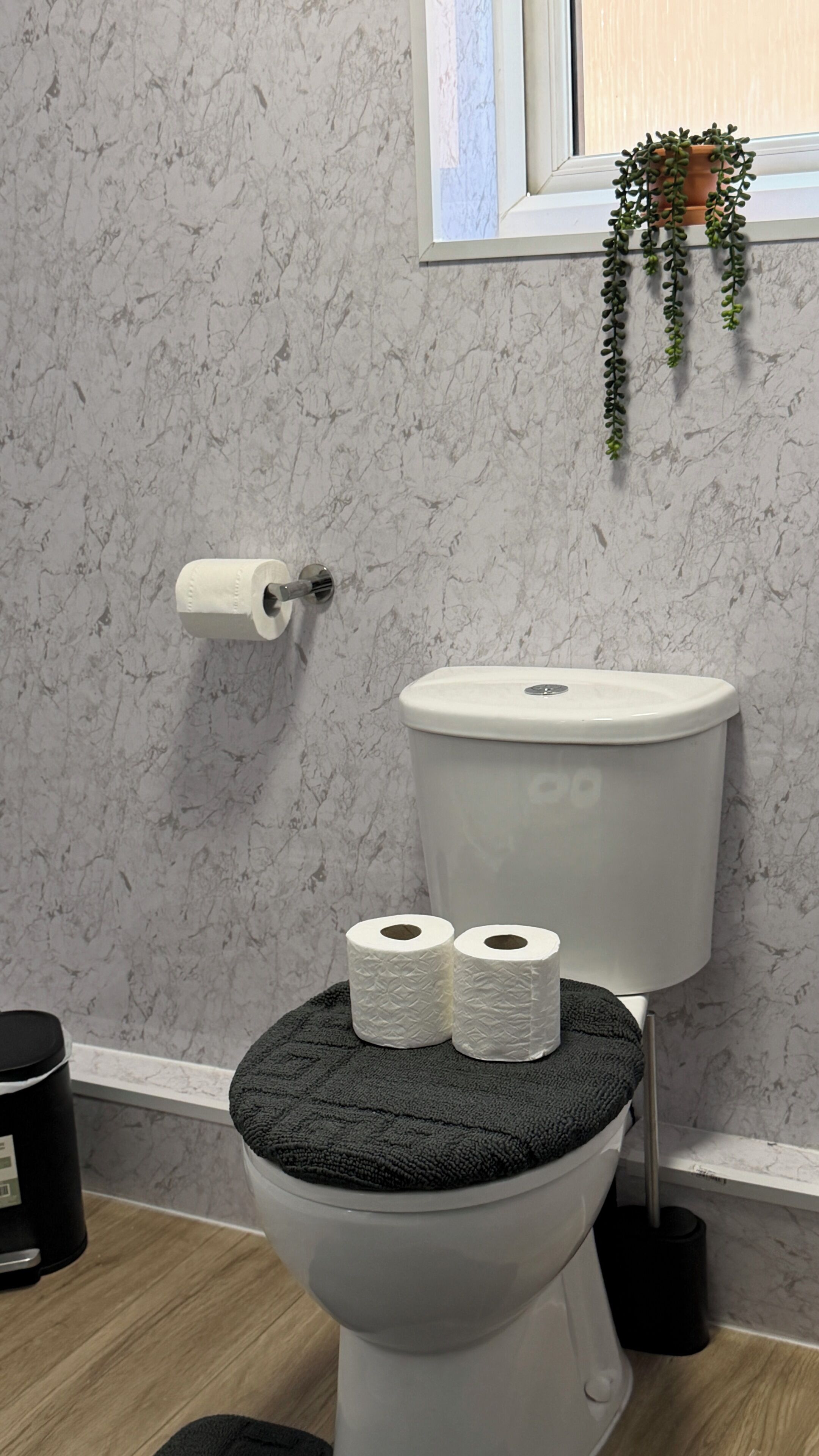 A toilet with two rolls of toilet paper, a black mat on the seat, a potted plant on the window sill, and a marble-patterned wall.
