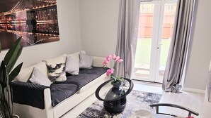 A modern living room with a sofa, a dining table set with plates and glasses, a potted plant, and a cityscape painting. - Modern 2BR 5minutes walk Anfeild| Free parking+ Ev (Liverpool)