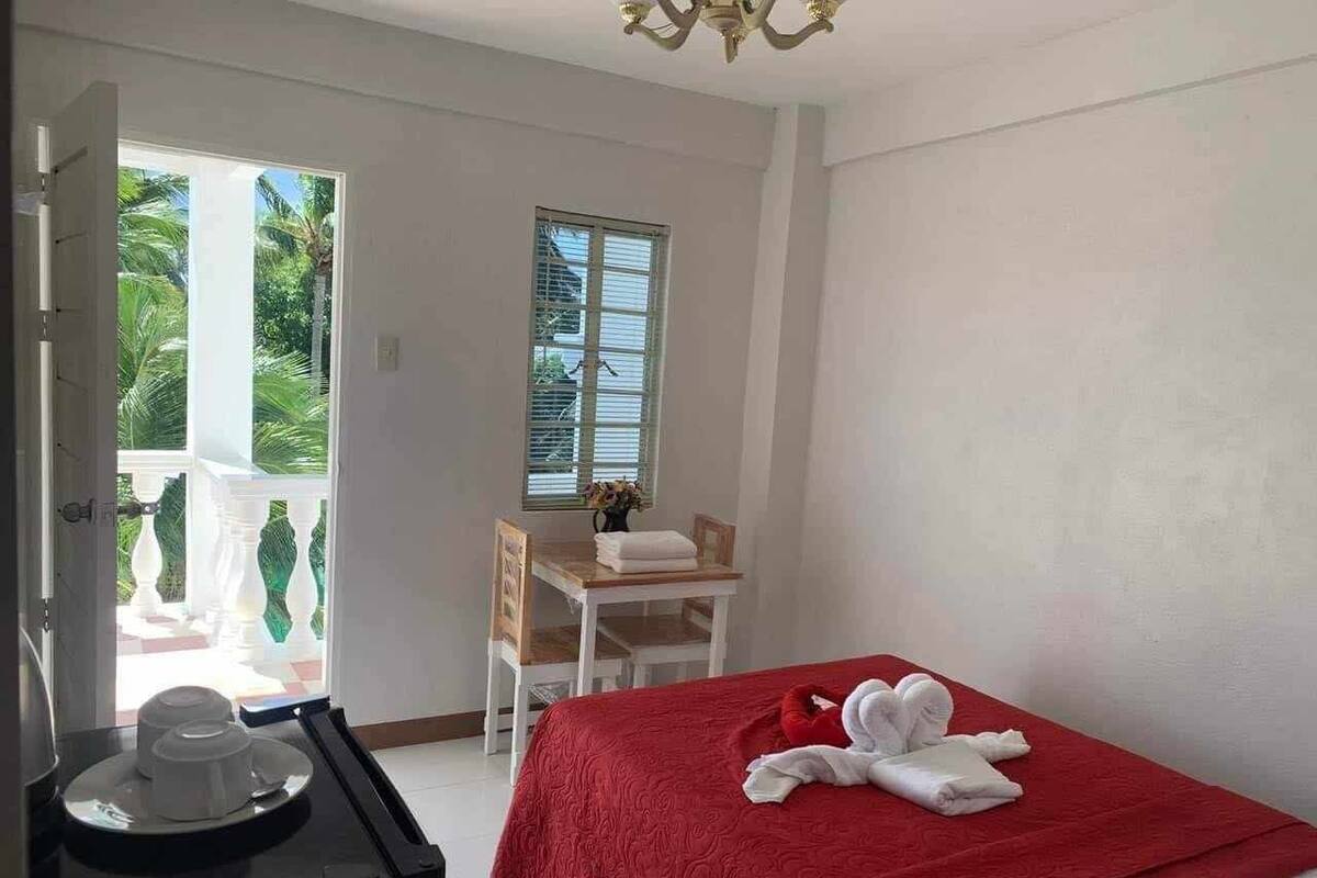 A hotel room with a bed, a table with a chair, a window, and a balcony with a view of greenery.