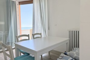 Une pièce avec une table blanche, deux chaises, une vue sur la mer par une fenêtre ornée de rideaux et un radiateur.