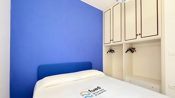 Un dormitorio con pared azul, cama blanca y armario con ropa colgada.