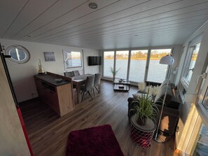A modern interior with a wooden ceiling, a dining area, a seating area with a view of the water, and a television mounted on the wall. - Modern, dog-friendly houseboat on Lake Wangermeer with a terrace, lake views, SUP, swimming access, and air conditioning—perfect for a relaxing vacation with family, friends, or as a couple. (Wangerland)